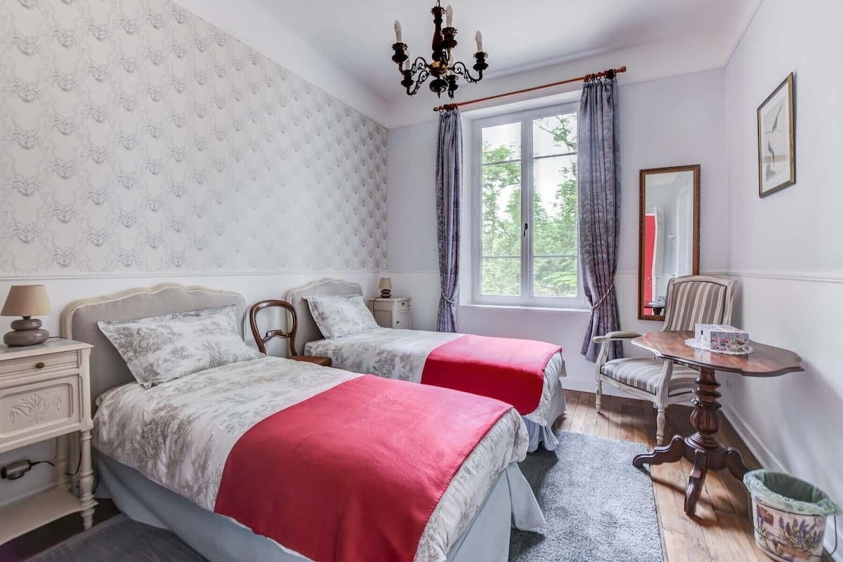Un dormitorio con dos camas, una silla de madera, una mesita, un espejo y una ventana con cortinas.