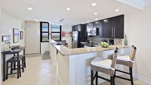 A modern kitchen with dark cabinets, a granite countertop, and a central island with bar stools. - Condo on Beach | Pool, Gym & Tennis Courts (Marco Island)