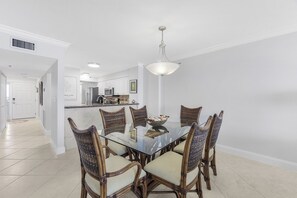 A dining area with a glass table surrounded by wicker chairs, a kitchen in the background, and a white interior.