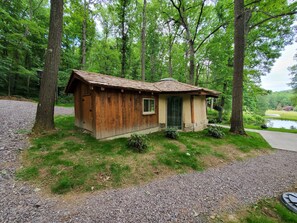 Una pequeña cabaña de madera con techo inclinado, rodeada de árboles y un camino de grava.