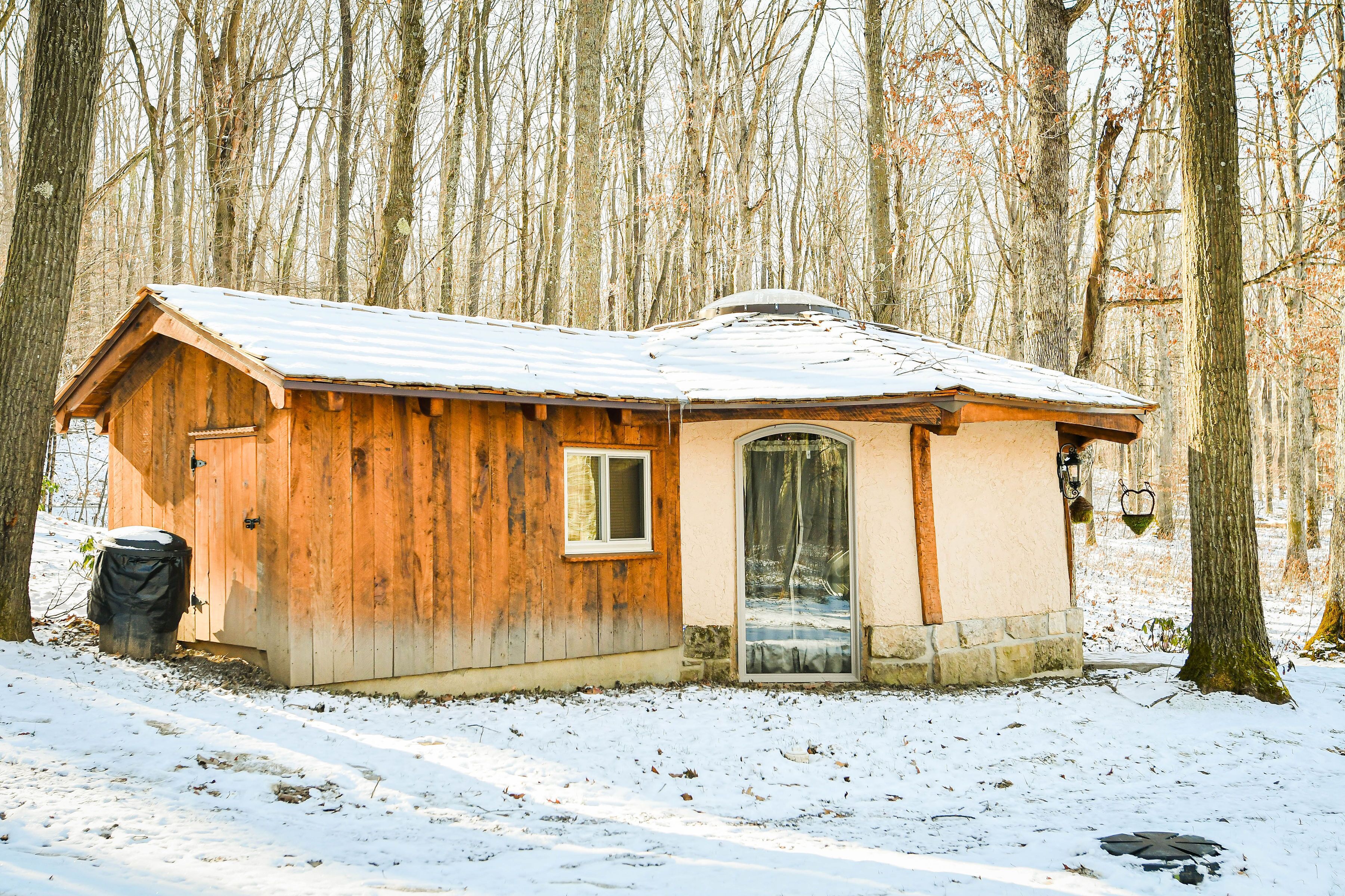A small wooden cabin with a metal roof, a door, and a window, surrounded by a snowy forest.