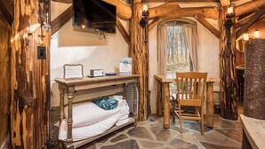 Ein rustikaler Raum mit Holzboden, einem Bett, einem Tisch, einem Stuhl, einem Wandfernseher und einem Fenster mit Vorhängen.
