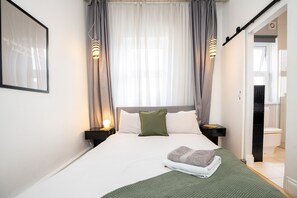 Un lit bien fait, recouvert d’une couverture verte, avec un oreiller et une serviette pliée. On y voit deux tables de chevet avec des lampes, un tableau encadré au mur et une salle de bain visible à travers une porte ouverte.