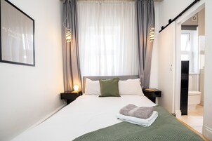 A neatly made bed with a green blanket, a pillow, and a folded towel. There are two bedside tables with lamps, a framed picture on the wall, and a bathroom visible through an open door.
