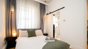 En snyggt bäddad säng med vita sängkläder, en grön pläd och en hopvikt handduk. Det finns två sänglampor, ett mörkt nattduksbord och en spegel på väggen.