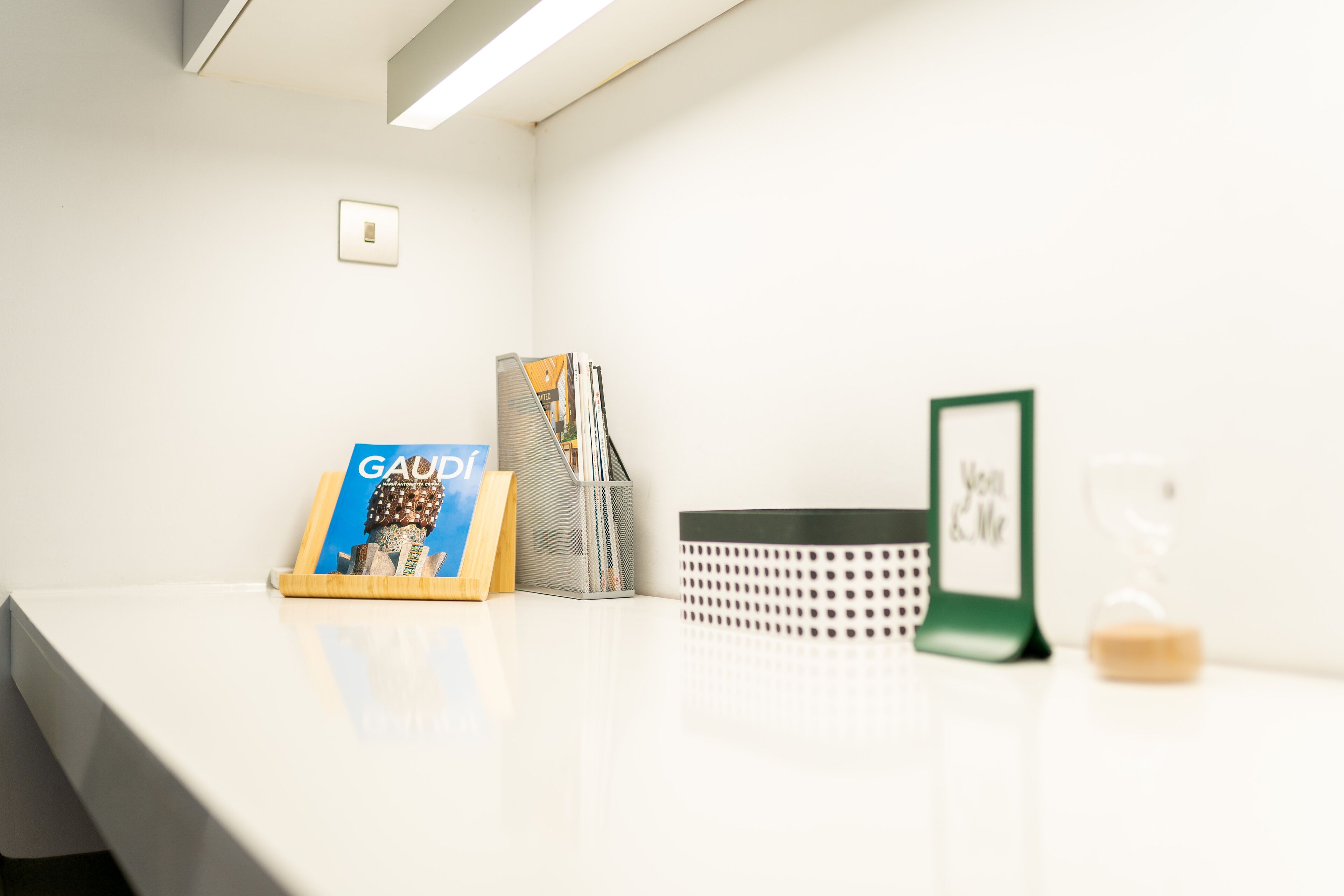 A clean, white desk with a framed picture, a book, a pen holder, a perforated basket, and an hourglass.