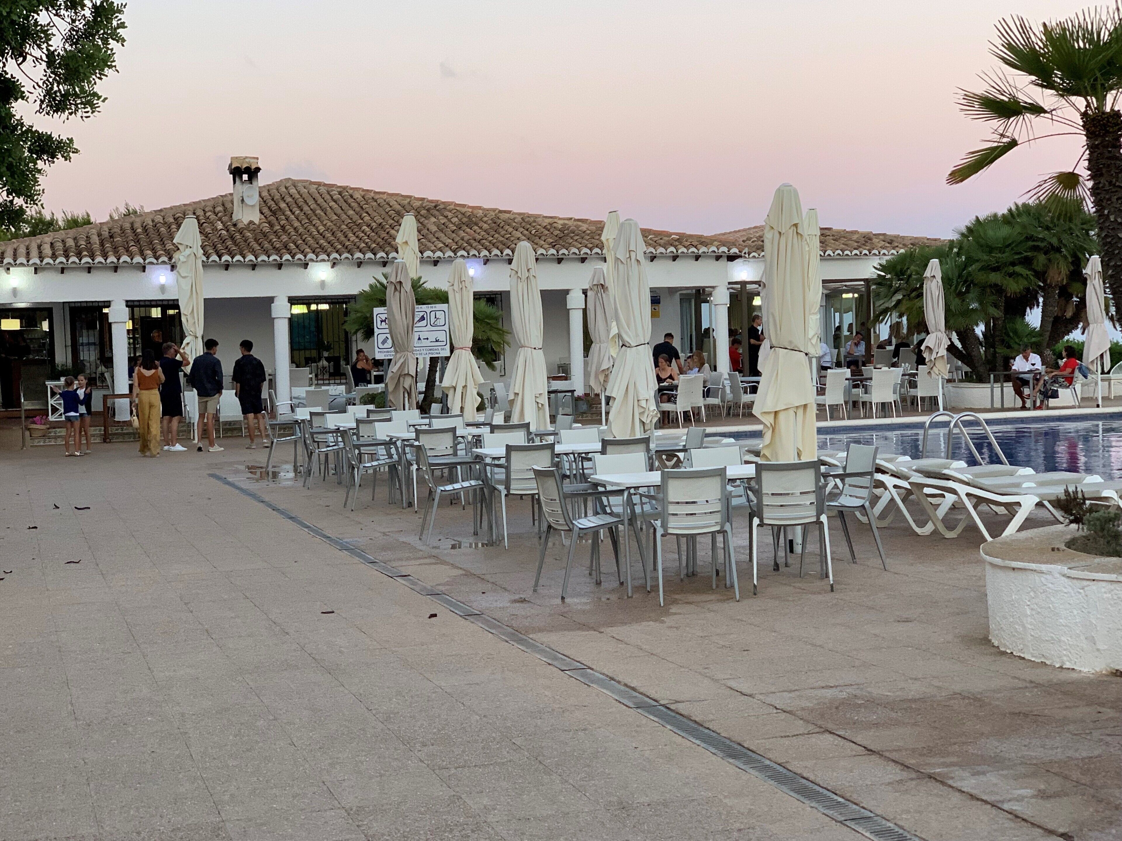 An outdoor cafe area with tables and chairs, umbrellas, and a pool in the background.