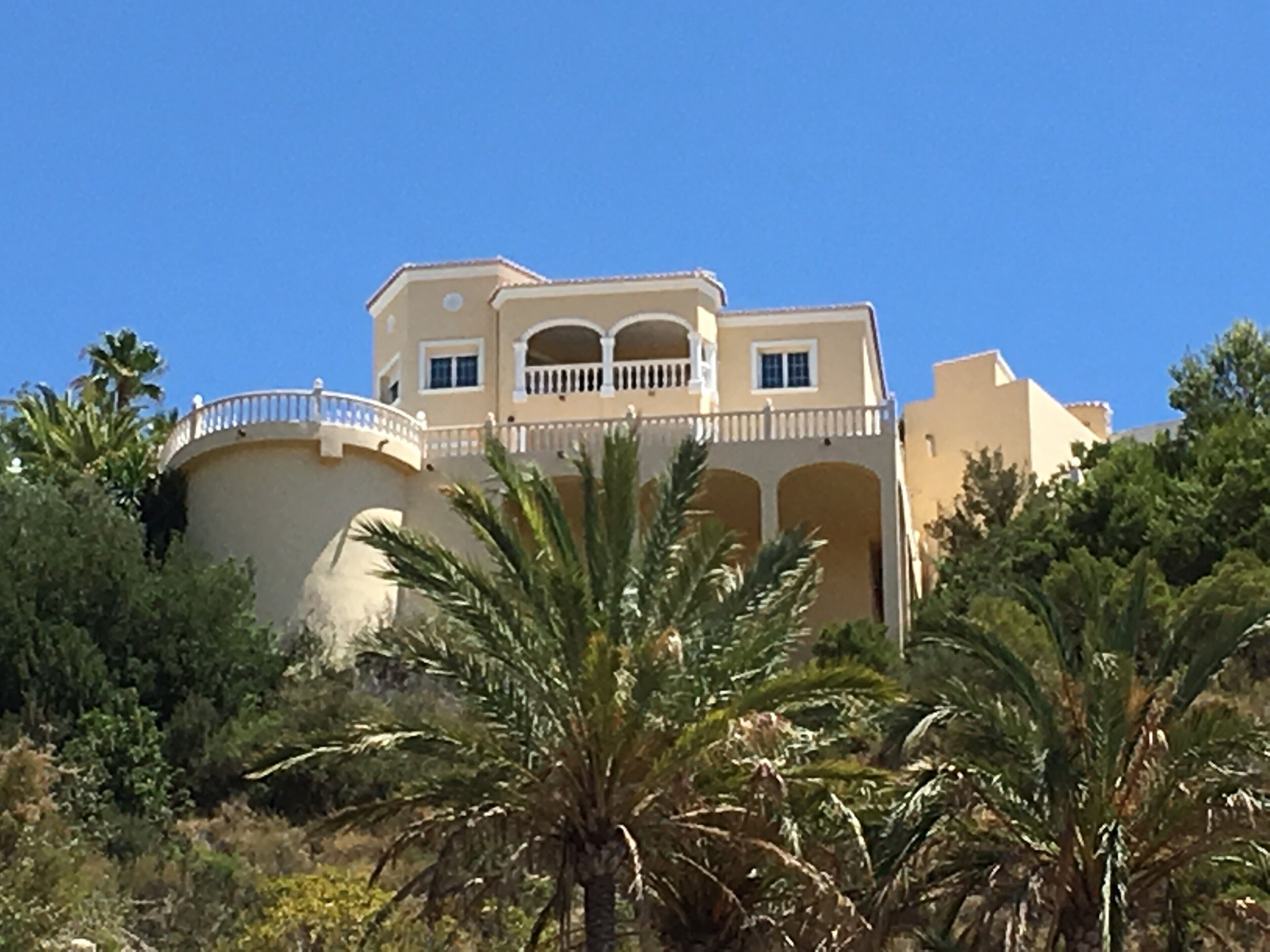 A large, multi-story building with a curved balcony, surrounded by palm trees and other vegetation.