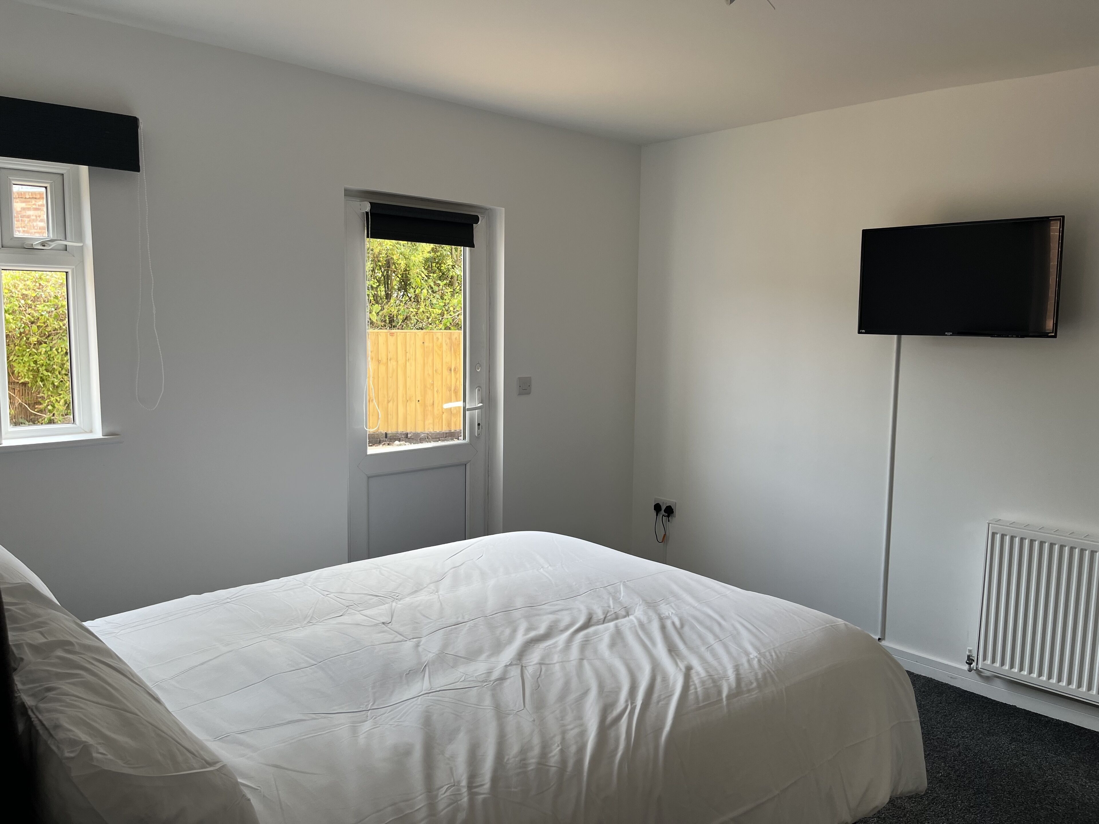 A bedroom with a bed, a window, a door, a wall-mounted TV, and a radiator.