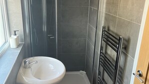 Un baño con un lavabo blanco, baldosas oscuras, cabina de ducha de vidrio y un perchero para toallas.