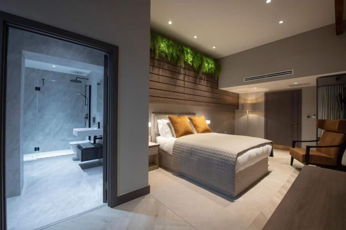 A modern bedroom with a bed, a nightstand, a chair, a built-in plant wall, and a bathroom with a shower and sink.