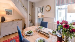 A dining area with a wooden table set for four, a clock on the wall, and a staircase leading to another room.