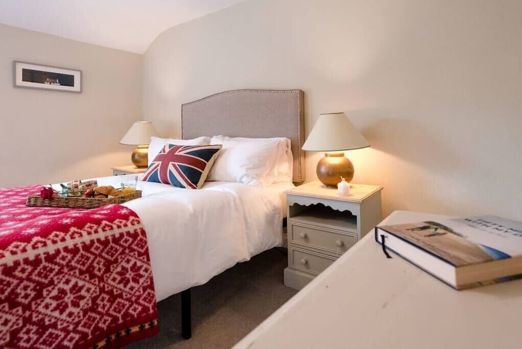 A neatly made bed with a red and white patterned blanket, a pillow with a Union Jack design, two bedside lamps, a bedside table with a book, and a framed picture on the wall.