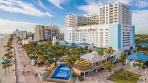 Um resort com vários edifícios altos, um calçadão à beira-mar e uma área de piscina central com guarda-sóis.