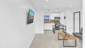 Uma sala de estar moderna com uma mesa de centro de madeira, um sofá cinzento e vista para uma cozinha com armários brancos e um espaço de jantar.