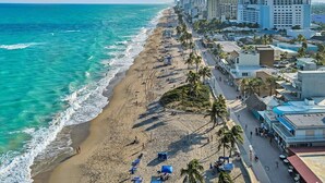 Paisagem costeira com praia, palmeiras e edifícios altos.
