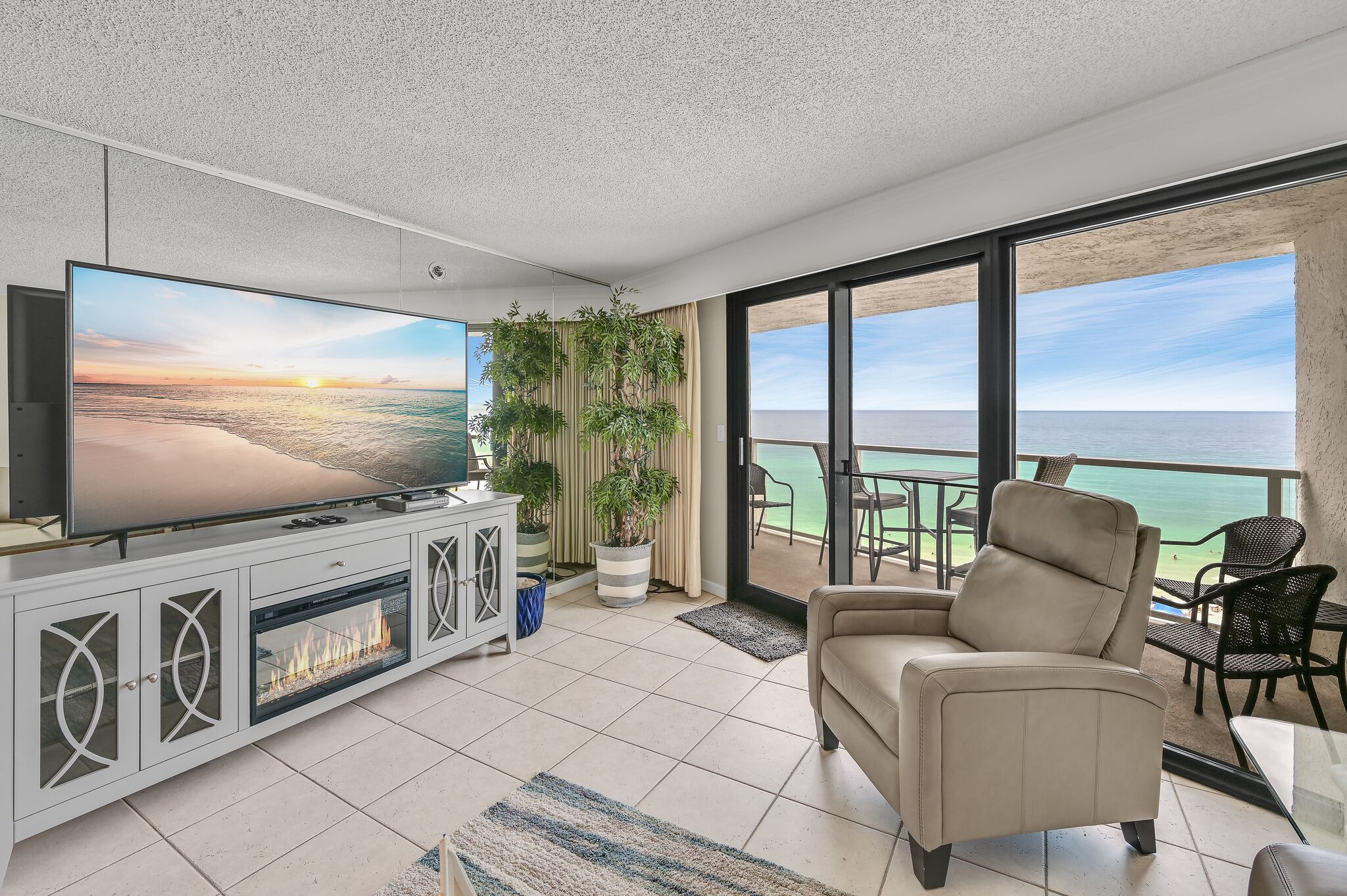 A modern living room with a large flat-screen TV, a beige sofa, and a balcony with a view of the ocean.