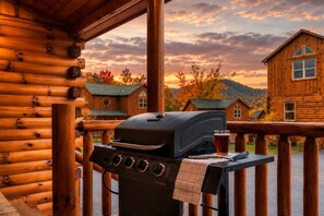 Una terraza de madera con una parrilla y una cerveza, con vistas a una hilera de cabañas de troncos y follaje otoñal.