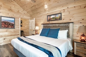 A wooden-paneled bedroom with a large bed, a nightstand with a lamp, and a framed picture on the wall.