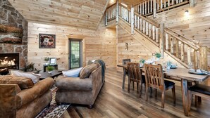 A cozy cabin interior with a stone fireplace, wooden walls, a staircase, a dining area, and a living room with sofas. - 1 Min to Parkway|4 King BRs|Hot Tub|Pool|Dogs (Pigeon Forge)