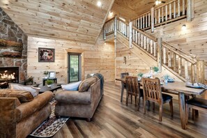 A cozy cabin interior with a stone fireplace, wooden walls, a staircase, a dining area, and a living room with sofas.
