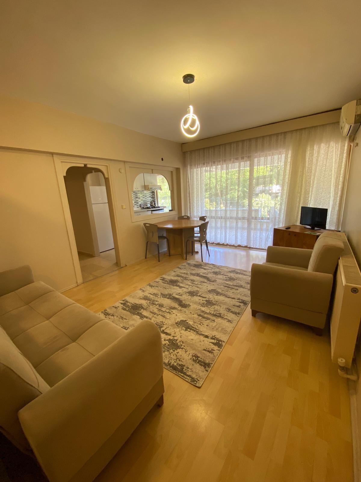 Un salon avec un canapé, une table basse et un téléviseur. On y trouve également un coin repas avec une table et des chaises, et une cuisine visible à travers une arche.