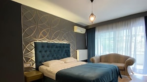 Un dormitorio moderno con cabecera capitonada, cama con cubrecama azul, mesita de noche, una silla y un ventanal con cortinas.