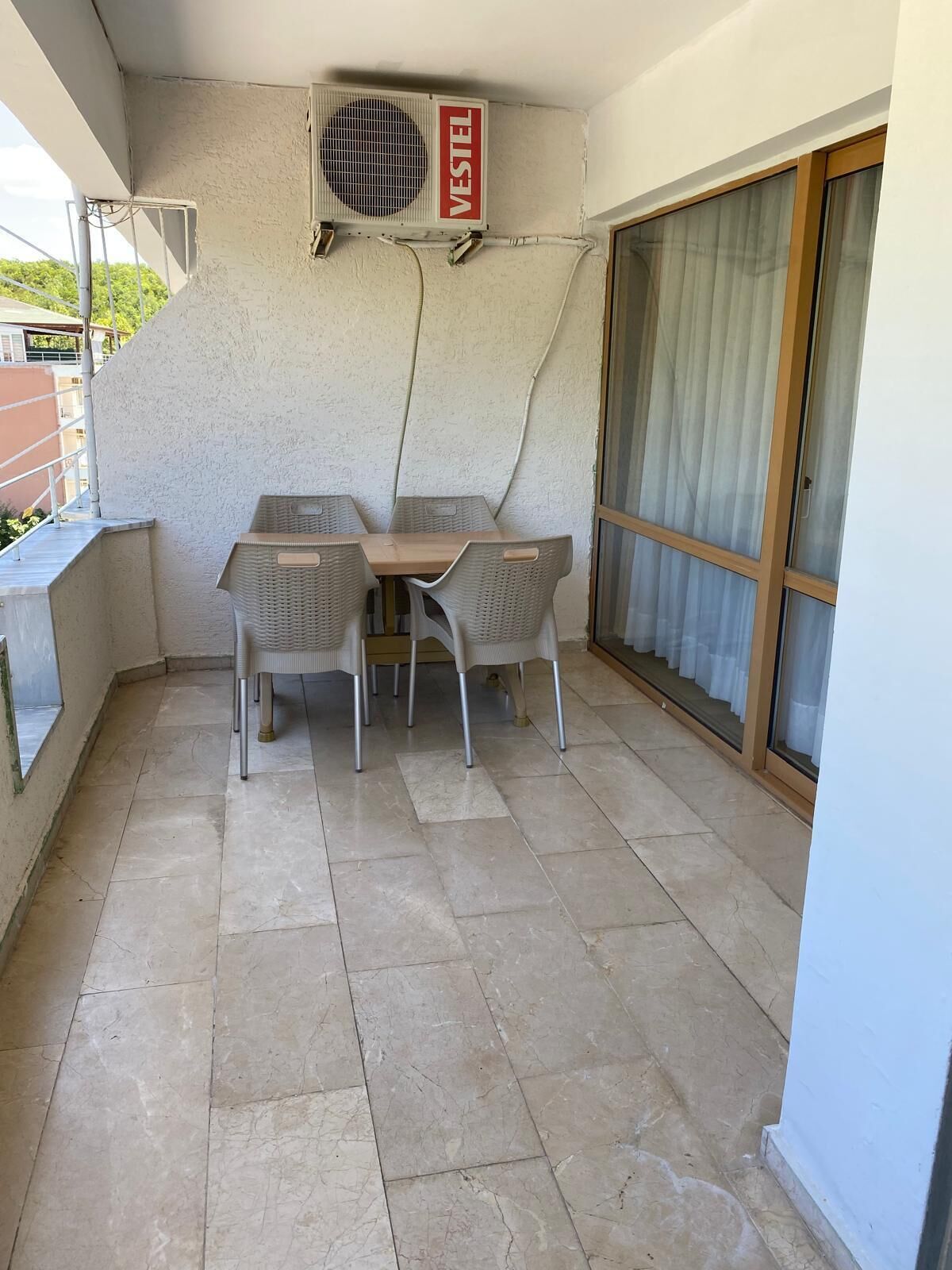 Un balcón con mesa y sillas, una puerta corredera de vidrio y un equipo de aire acondicionado con la etiqueta 'VESTEL'.