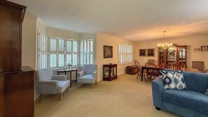 A living room with a blue sofa, striped armchairs, a wooden coffee table, and a dining area with a chandelier.