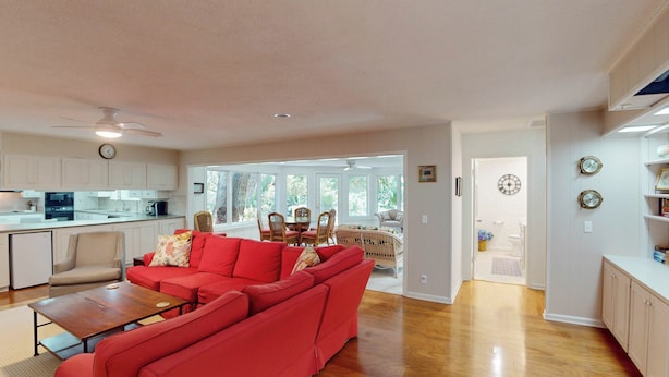 A spacious living room with a red sectional sofa, a wooden coffee table, and a dining area with wicker chairs. A kitchen with white cabinets and a large window is visible in the background. - Lilly's Pad 3 bedrooms, 3.5 bathrooms (Jekyll Island)