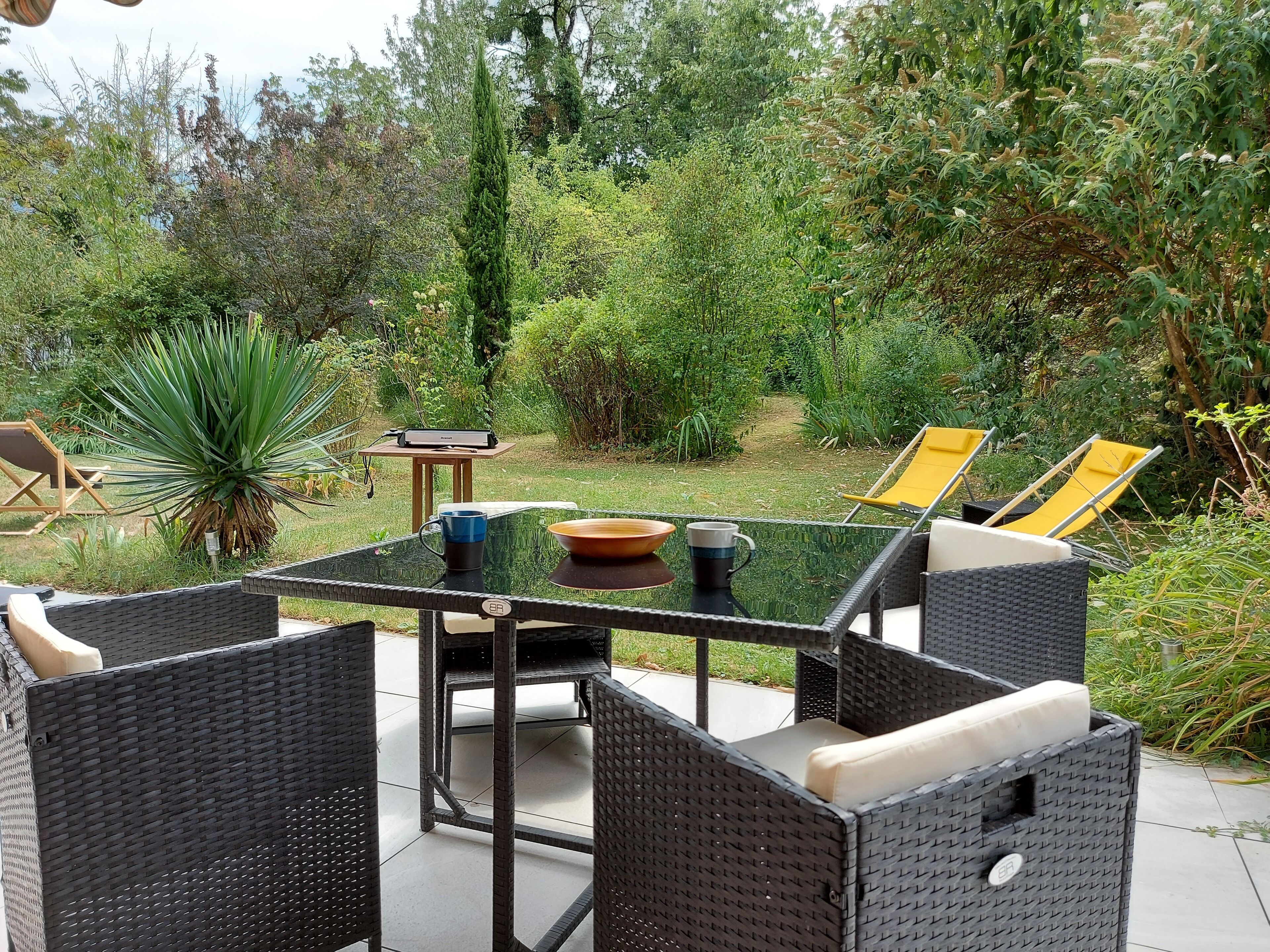 An outdoor patio with wicker furniture, a glass table, and a view of a garden.