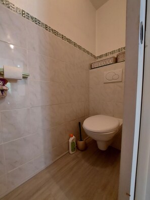 Casa de banho com sanita, secador de mãos, suporte de papel higiénico e frasco de produto de limpeza.