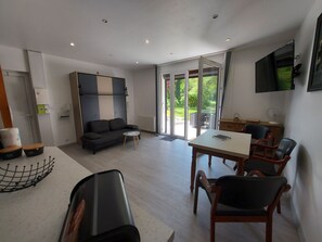 A modern living room with a sofa, a dining table with chairs, a TV mounted on the wall, and a sliding glass door leading to an outdoor area. - Centre St Jorioz : Charmant et Spacieux Studio Avec Terrasse et Jardin Privatif (Saint-Jorioz)