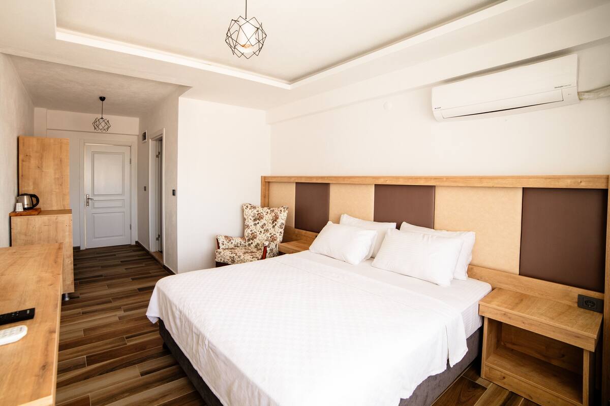 A hotel room with a large bed, wooden headboard, a small wooden nightstand, a chair with a floral cushion, a wooden desk with a kettle, and an air conditioning unit on the wall.