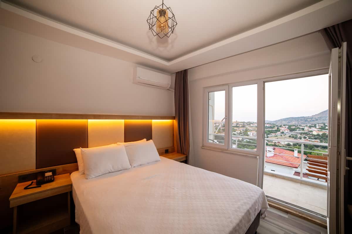 A hotel room with a bed, bedside table, and a view of the city through a large window.