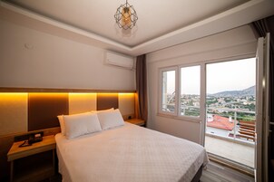 Habitación de hotel con cama, mesita de noche y vistas a la ciudad a través de un gran ventanal.