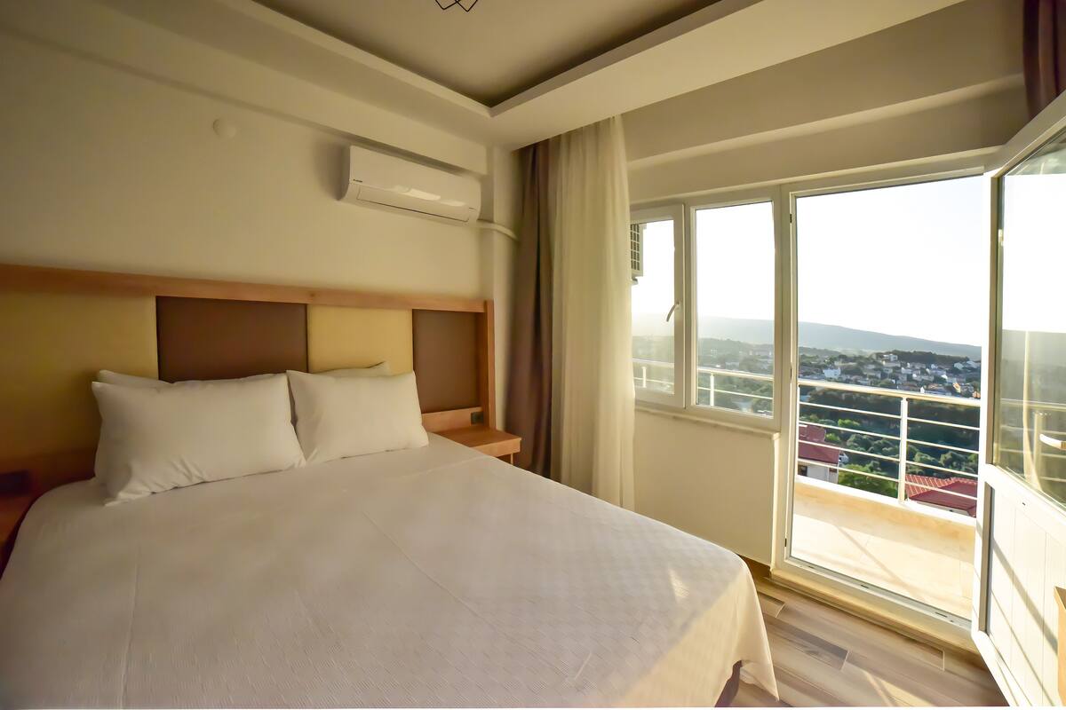 A hotel room with a large bed, an air conditioning unit, a wooden headboard, a nightstand, and a large window with a view of a cityscape.