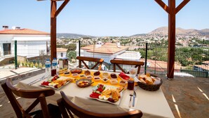 Una mesa dispuesta con diversos alimentos y bebidas, un pérgola de madera, y vistas a un pueblo y montañas.