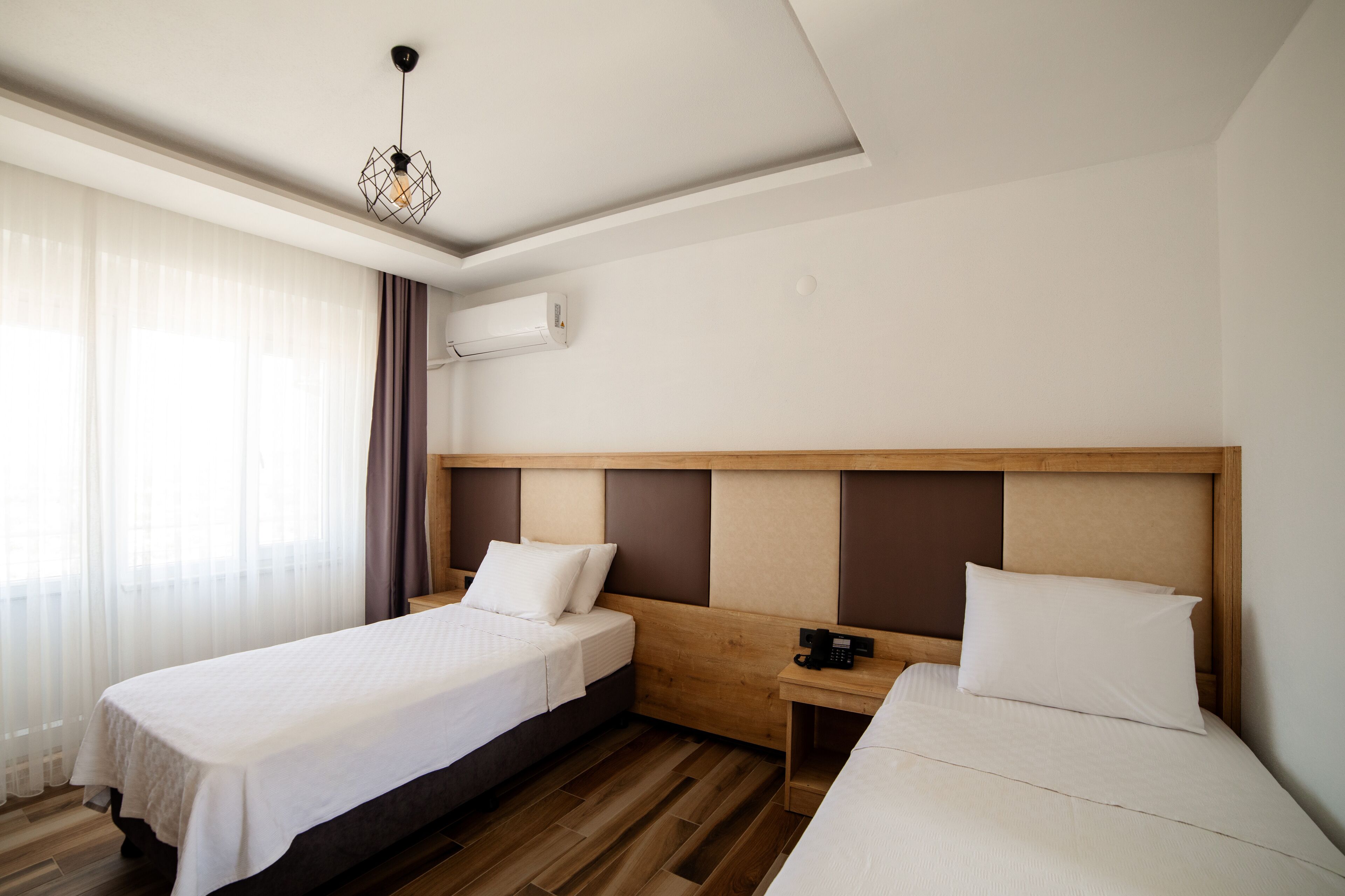 Une chambre d’hôtel avec deux lits simples, une tête de lit en bois, une table de chevet avec un téléphone et une fenêtre avec des rideaux légers.