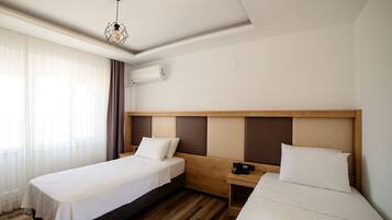 Une chambre d’hôtel avec deux lits simples, une tête de lit en bois, une table de chevet avec un téléphone et une fenêtre avec des rideaux légers.