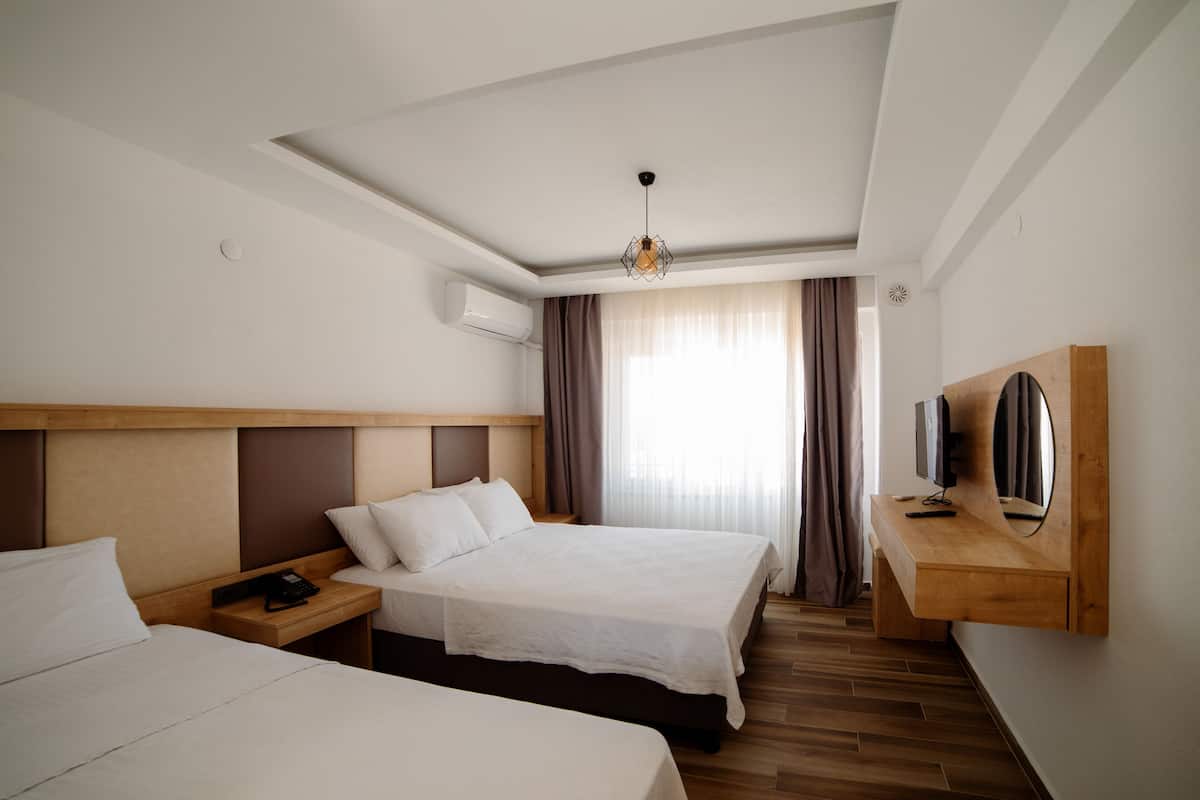 A hotel room with two beds, a wooden headboard, a bedside table with a phone, a TV mounted on a wooden panel, and a window with curtains.