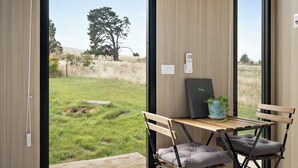 A room with a view of a grassy field and a tree, a desk with a laptop, and a chair with a cushion. - Tiny Regen Farm Stay by Tiny Away (Lawrence)