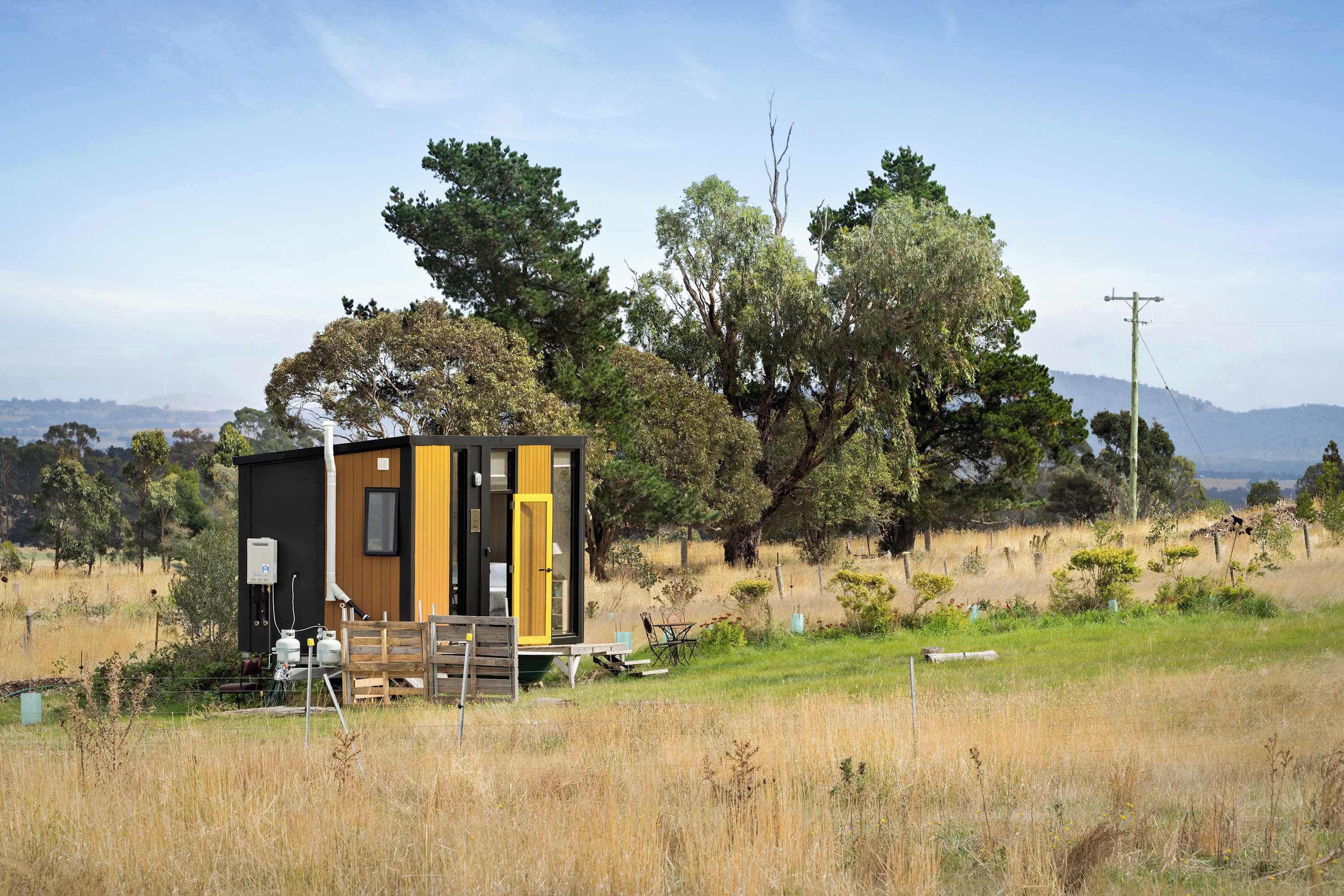 A small cabin with a wooden exterior, a black door, and a yellow window, surrounded by trees and a grassy field.