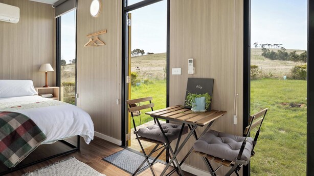 A bedroom with a bed, bedside table, lamp, and a view of a grassy field through a large window. - Tiny Regen Farm Stay by Tiny Away (Lawrence)