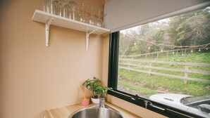 Eine kompakte Küche mit einer Spüle aus Edelstahl, einem Fenster mit Blick auf eine grüne Landschaft und einem Regal mit Gläsern.