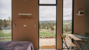 Ein Zimmer mit einem Bett, einem Tisch und einer Bank. Durch ein großes Fenster ist ein Blick auf eine Wiese und Bäume möglich.
