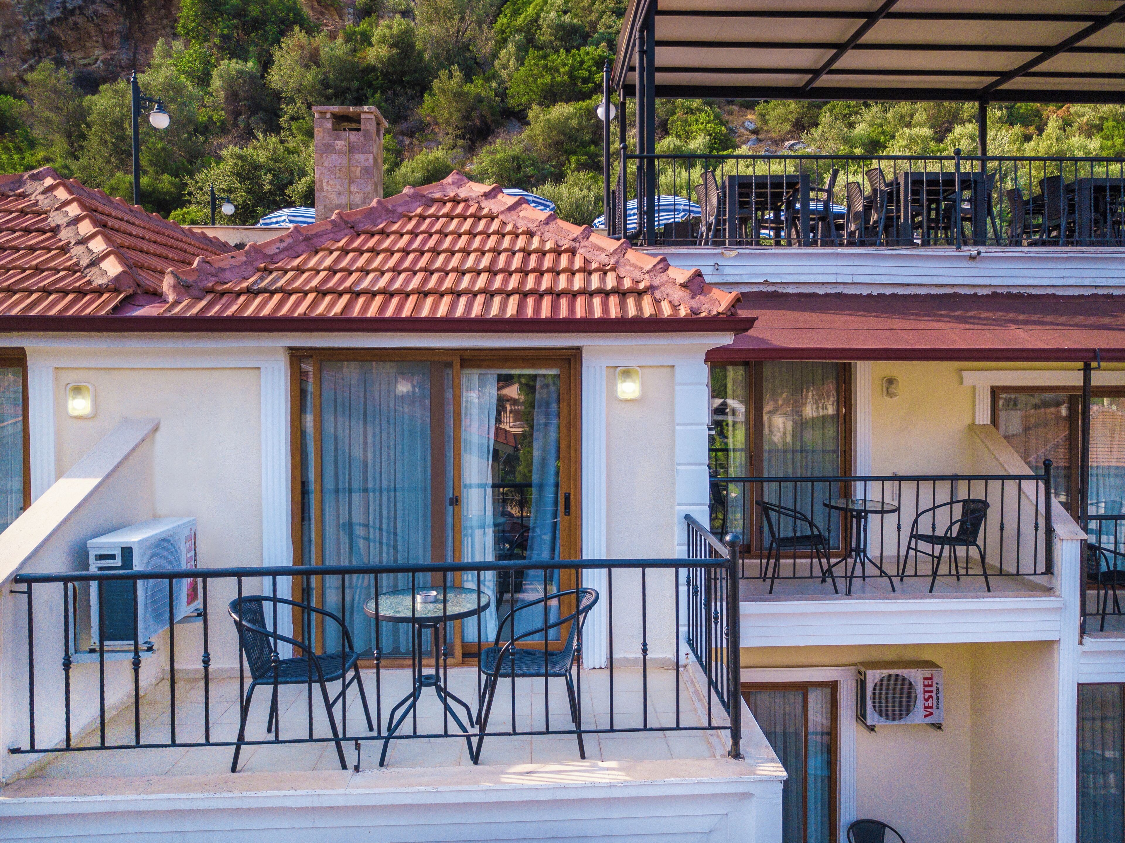A two-story building with a red-tiled roof, a balcony with outdoor seating, and a view of the surrounding greenery.
