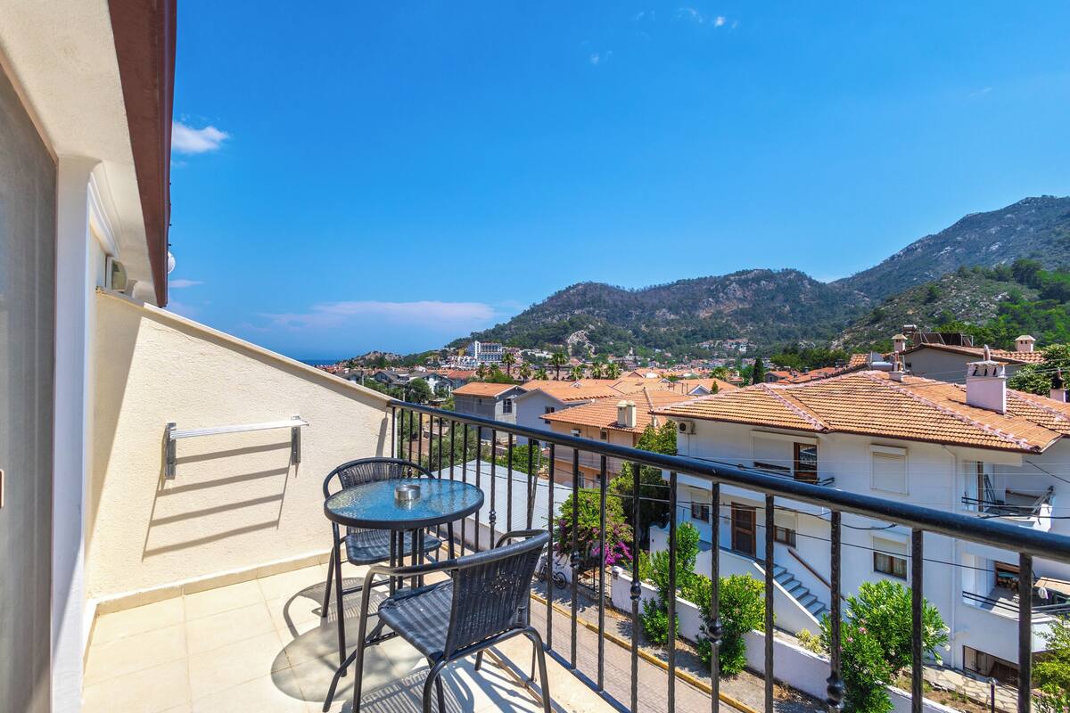A balcony with a table and chairs, overlooking a town with buildings and mountains in the background.