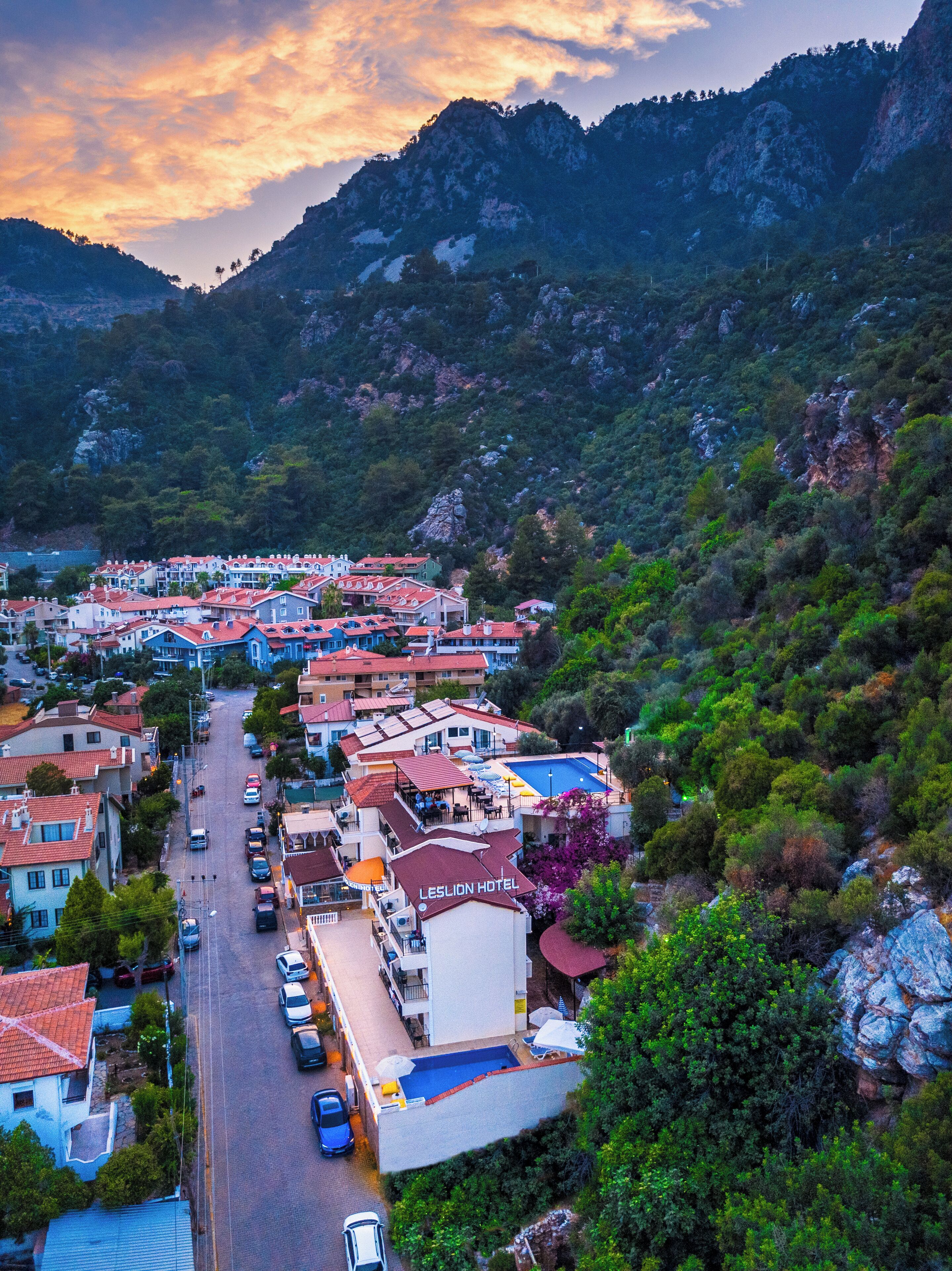 Un pueblo en la ladera de una colina con un hotel destacado llamado "LESLION HOTEL", que cuenta con una piscina en la azotea y vistas a las montañas circundantes.