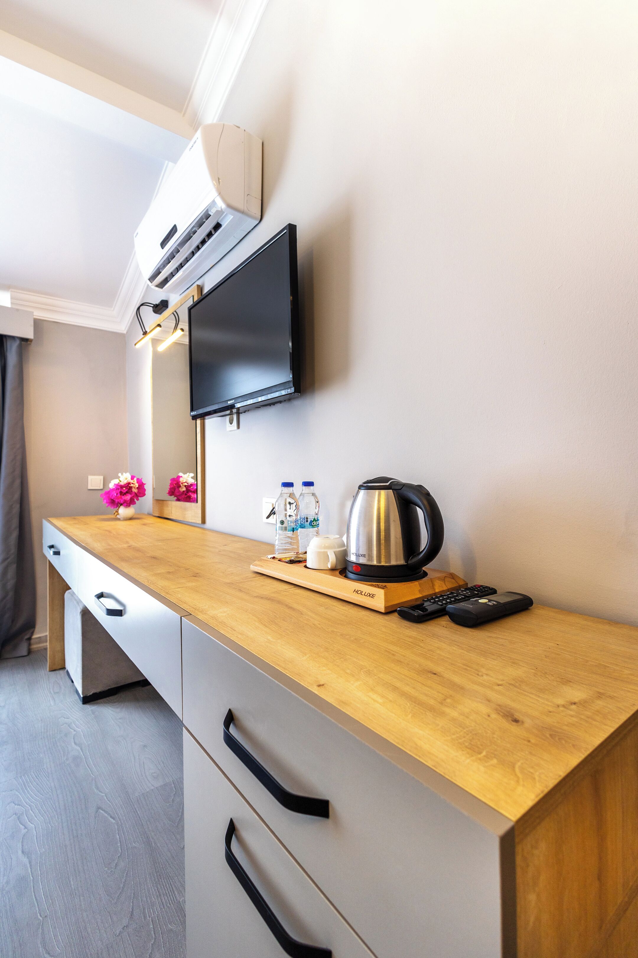 A modern hotel room with a wooden desk, a flat-screen TV mounted on the wall, a kettle, a water bottle, and a remote control.